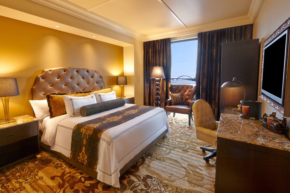 A hotel room with a large bed, two bedside tables with lamps, a desk with a chair, a large window with a view of a bridge, and a TV on the desk.