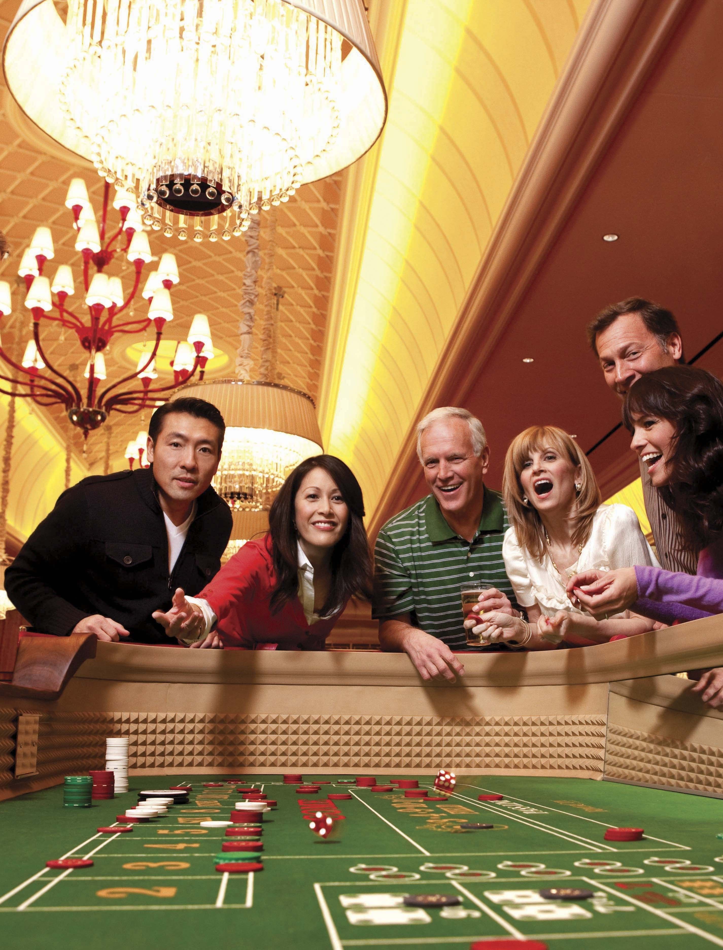 A group of people at a craps table in a casino with a chandelier and a high ceiling.