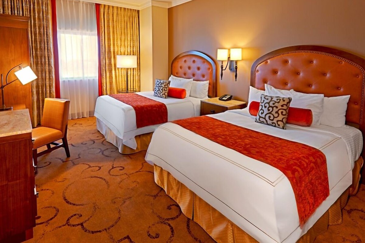 A hotel room with two beds, a desk, a chair, a lamp, and a window with curtains.