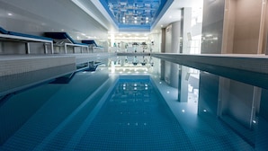 Ein moderner, überdachter Schwimmbereich mit klaren blauen Fliesen, einer Glasbarriere und einer Decke mit geometrischen Mustern.