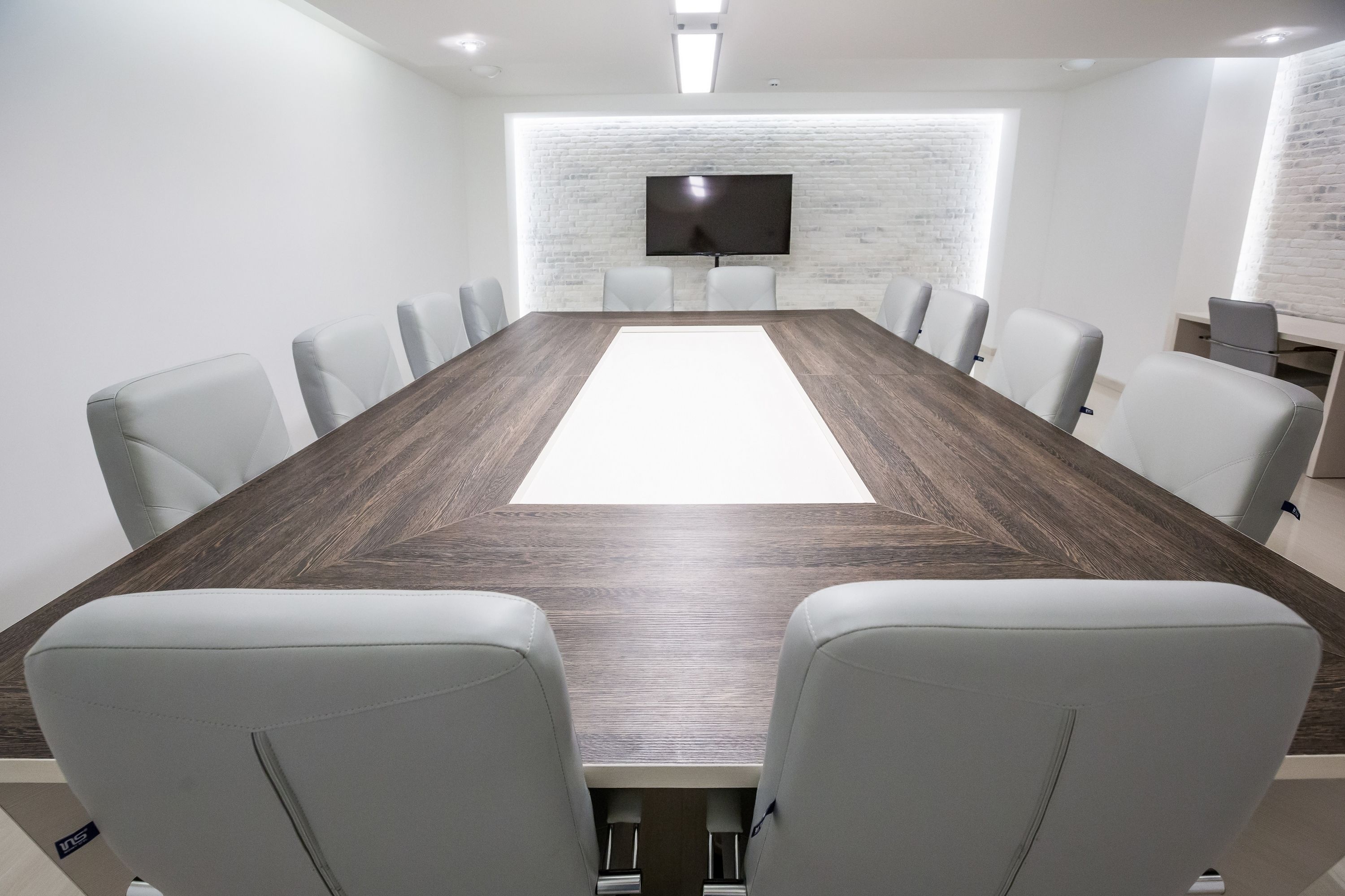 Una sala de conferencias con una mesa larga, sillas blancas, un televisor de pantalla plana en una pared de ladrillo y un escritorio pequeño con una computadora.