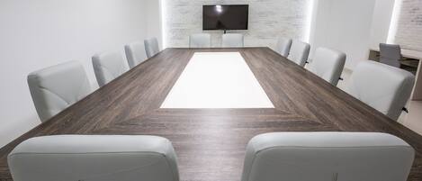 Una sala de conferencias con una mesa larga, sillas blancas, un televisor de pantalla plana en una pared de ladrillo y un escritorio pequeño con una computadora.