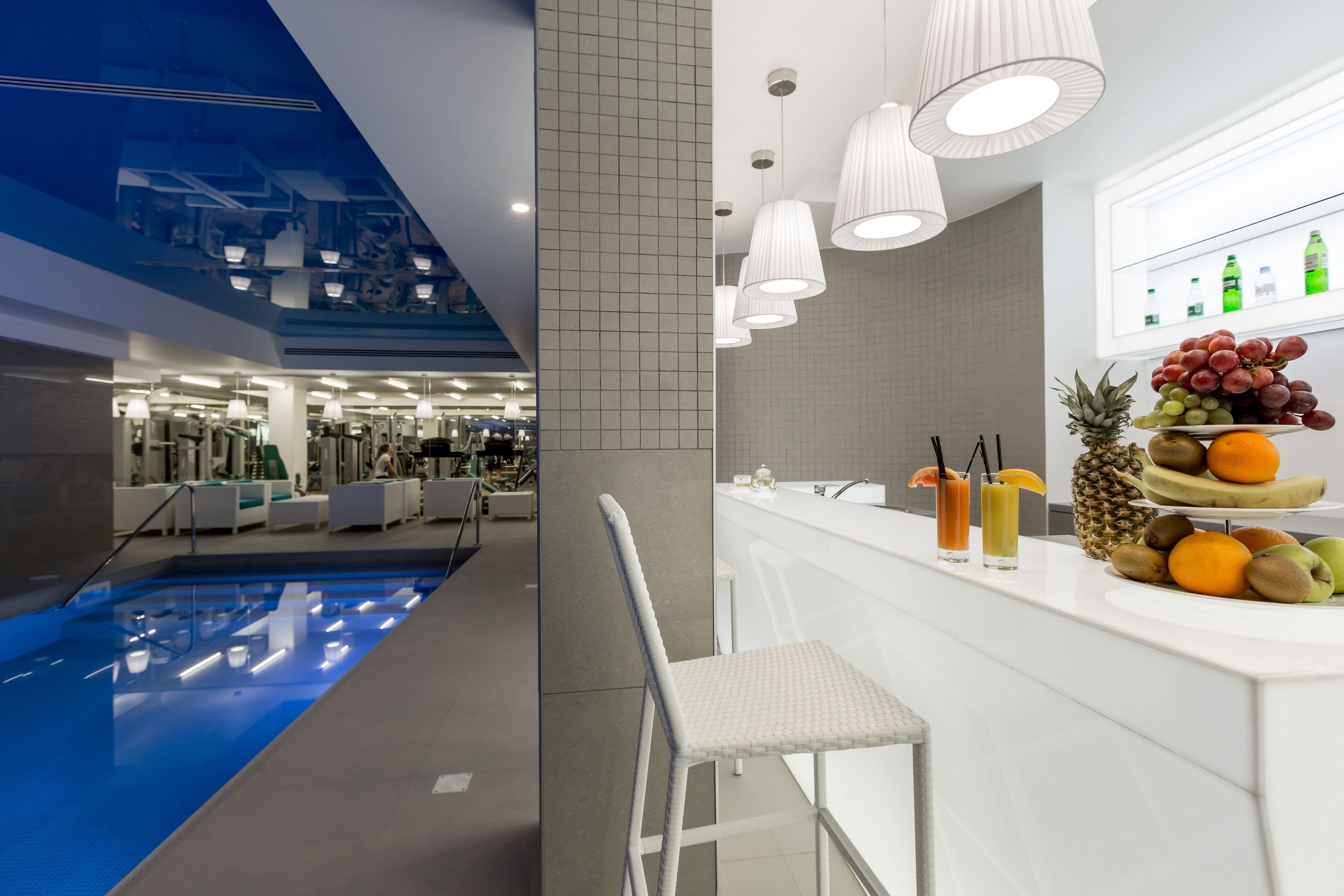 Un moderno vestíbulo de hotel con piscina, una zona de bar con frutas y bebidas, y vistas al área del gimnasio.