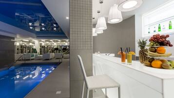 Un moderno vestíbulo de hotel con piscina, una zona de bar con frutas y bebidas, y vistas al área del gimnasio.