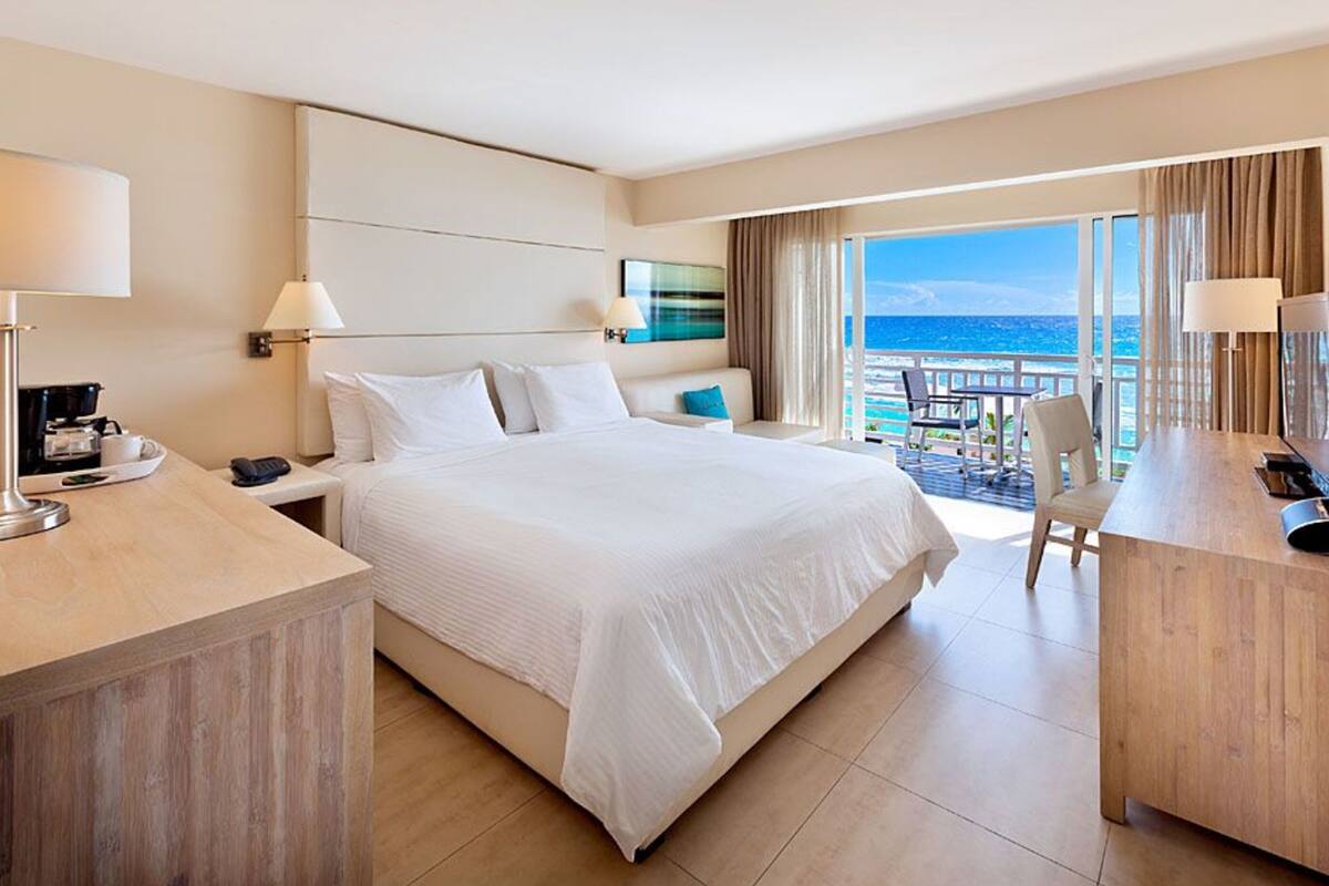 Quarto de hotel com uma cama grande, uma mesa com abajur, uma televisão e varanda com vista para o mar.