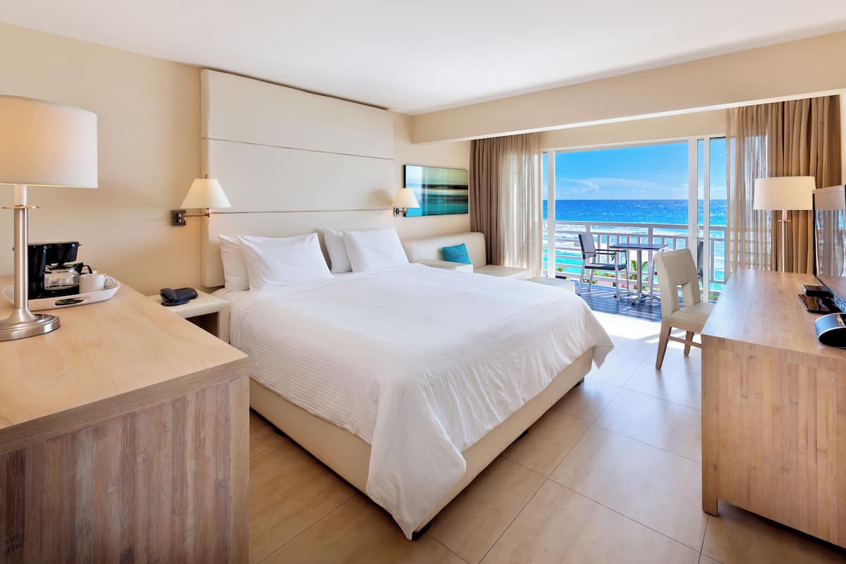 Quarto de hotel com uma cama grande, uma escrivaninha, uma cadeira e varanda com vista para o oceano.