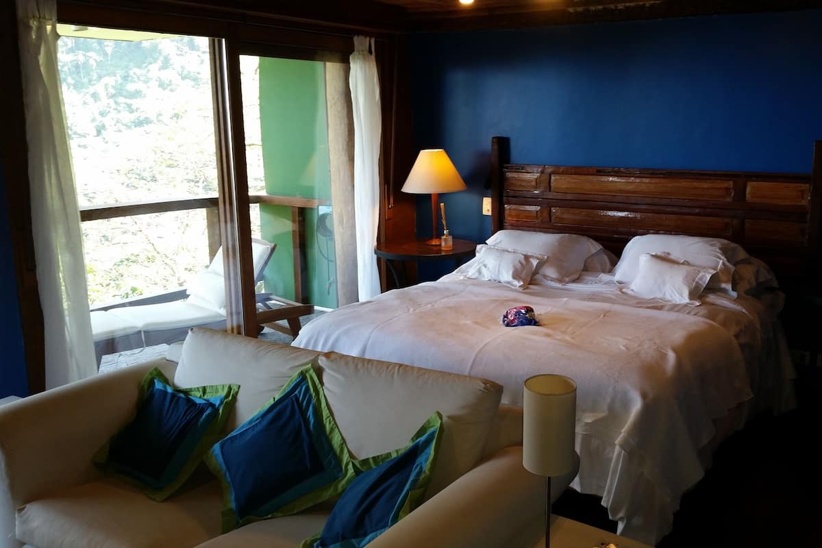 A bedroom with a bed, two bedside tables with lamps, a chair with cushions, and a view through a window.