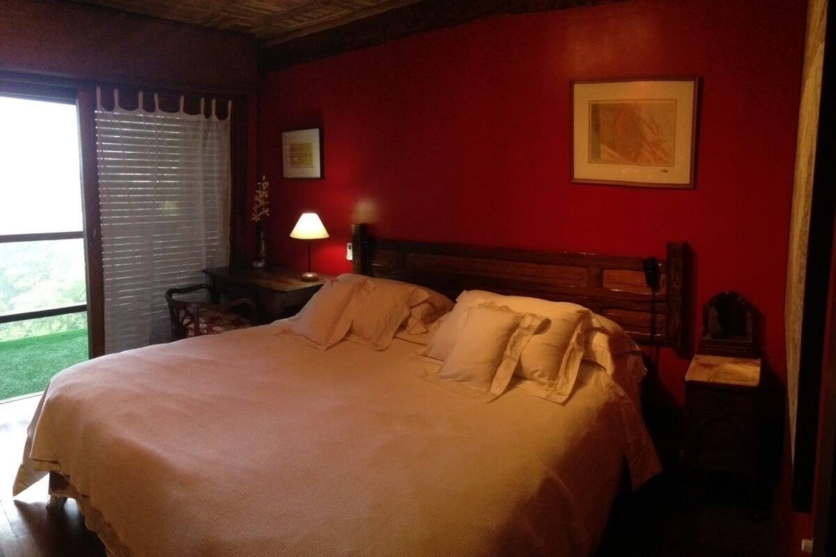 A neatly made bed in a room with red walls, a wooden headboard, and a window with blinds.