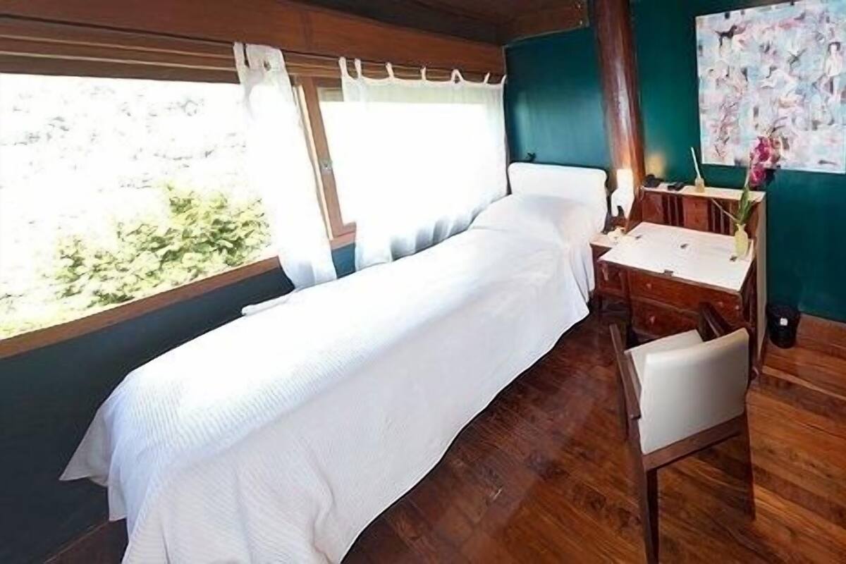 A single bed with a white bedspread, a wooden desk with a chair, and two abstract paintings on teal walls.