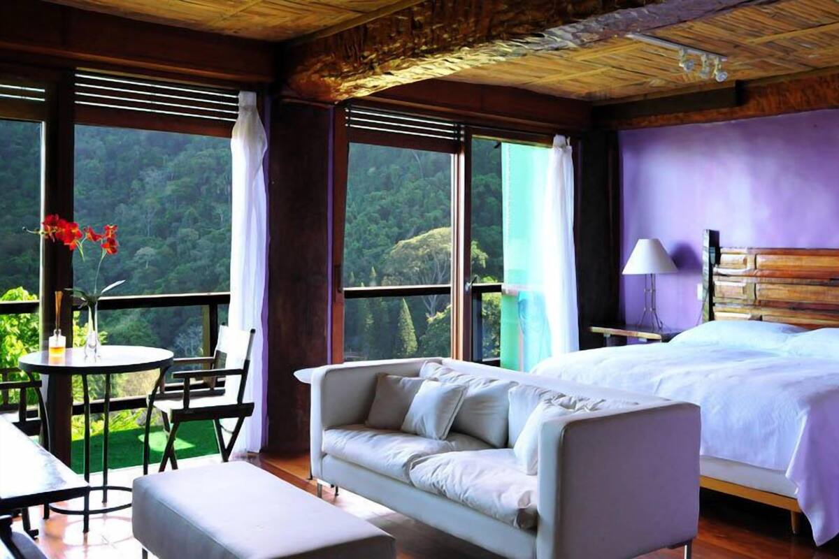 A bedroom with a bed, a sofa, a table, and chairs. There is a large window with a view of a forest.