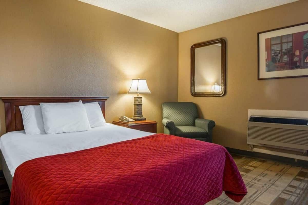 Ett hotellrum med en säng, en stol, en lampa, en spegel och en luftkonditioneringsanläggning.