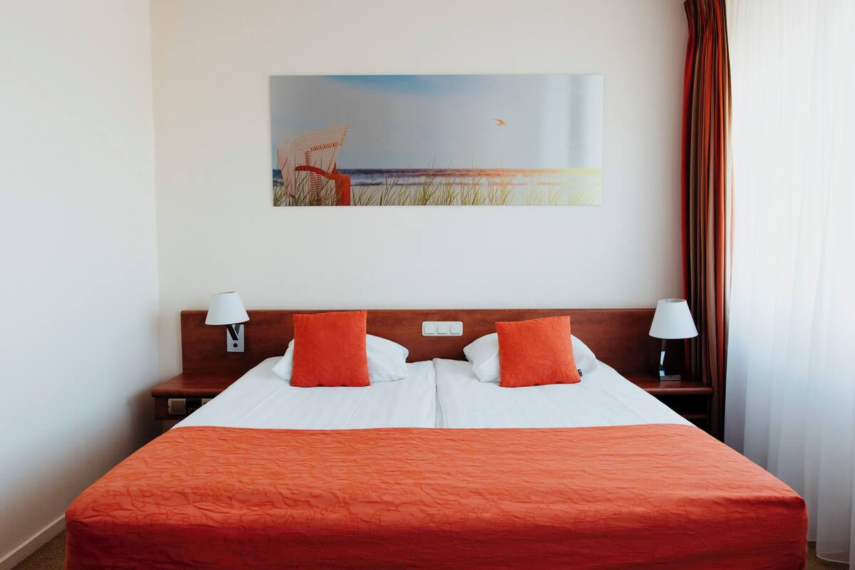 A hotel room with a large bed, two pillows, a nightstand with a lamp, a painting of a beach scene, and a window with curtains.