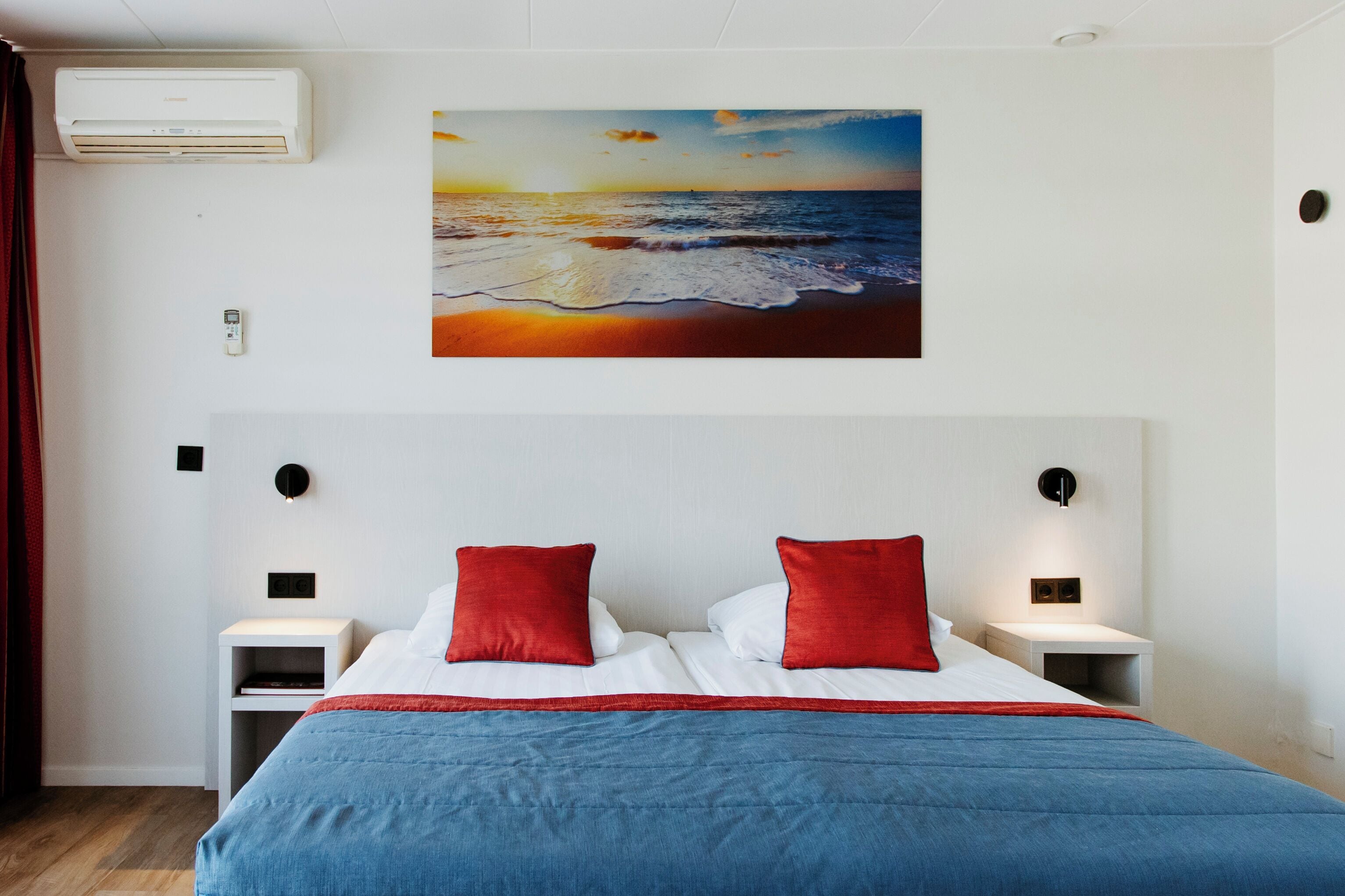 Une chambre à coucher avec un lit, des oreillers rouges, une couvre-lit bleue, un tableau représentant un coucher de soleil sur l’océan et un climatiseur blanc.