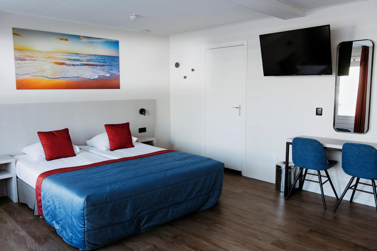 A hotel room with a bed, a desk, a chair, a TV, and a painting of a beach scene.