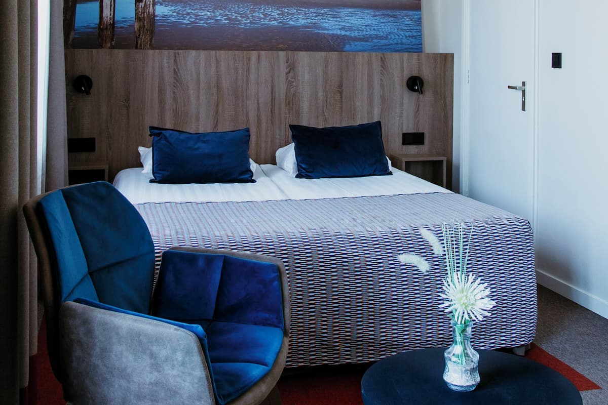 A hotel room with a bed, a chair, a small table, and a painting of a beach scene.