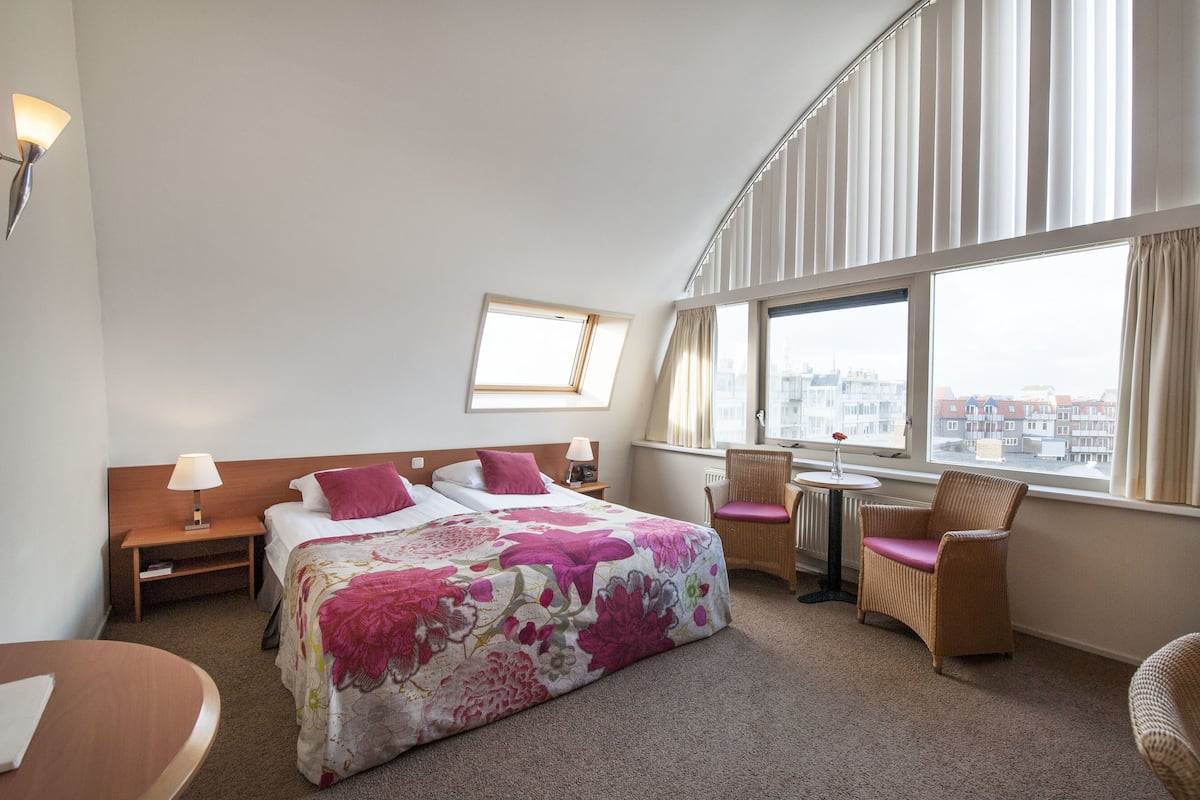 A hotel room with a bed, a desk, two chairs, a window with curtains, and a nightstand with a lamp.