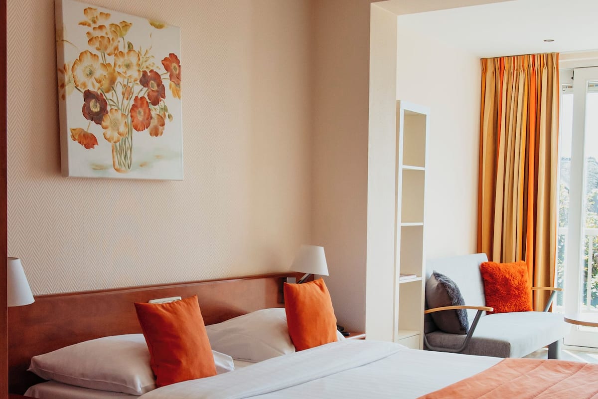 A hotel room with a large bed, two pillows, a nightstand, a chair, a window with curtains, and a painting on the wall.