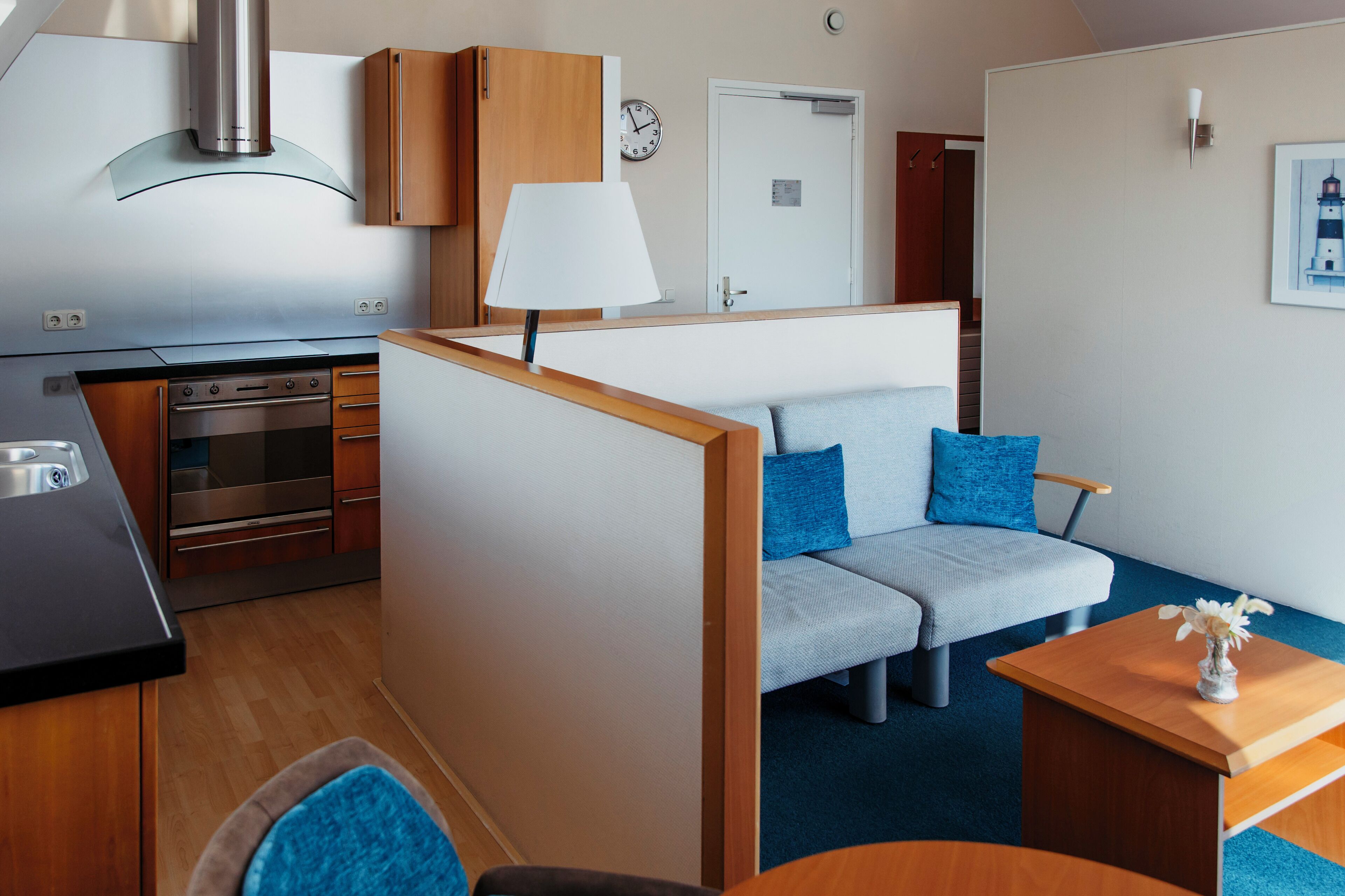 A compact living space with a kitchenette, a sofa with blue cushions, a small table with a vase, and a door with a clock.