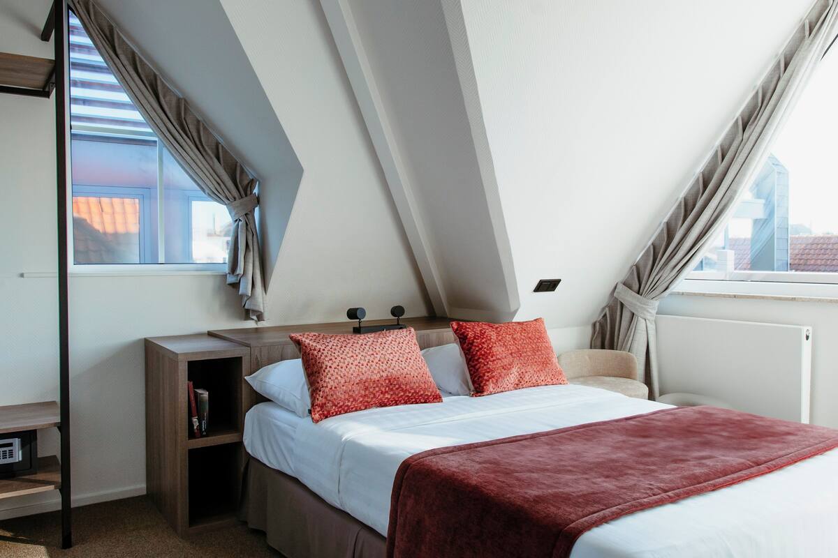 A bedroom with a sloped ceiling, a bed with white bedding and red pillows, a wooden nightstand, and a view of the city through large windows.