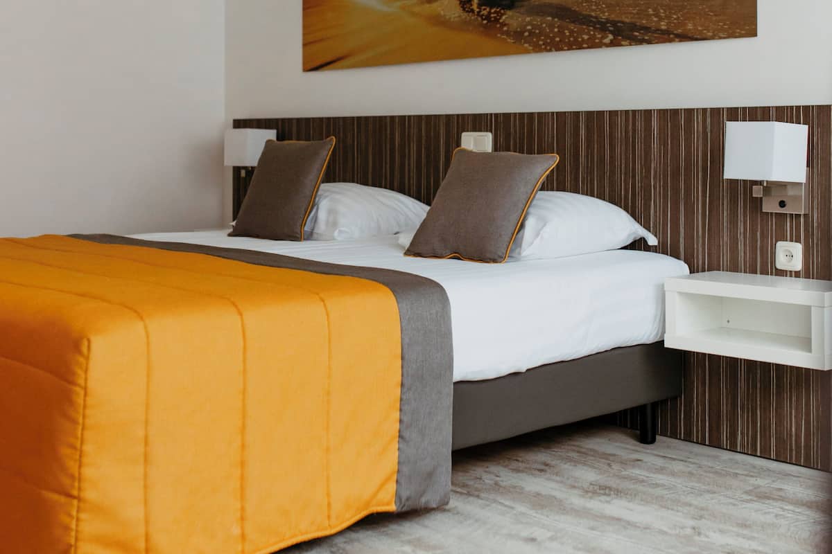 A modern hotel room with a large bed, a headboard with vertical lines, a bedside table with a lamp, and a painting of a sunset over the ocean on the wall.