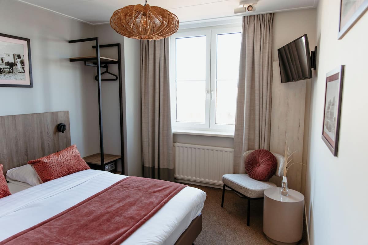 A bedroom with a bed, a chair, a side table, a wall-mounted TV, and a window with curtains.