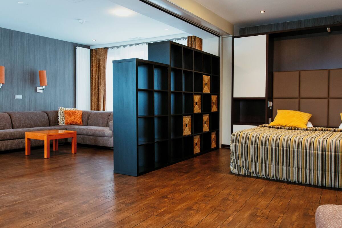 A modern hotel room with a wooden floor, a grey sofa with orange cushions, a striped bed, and a large black shelving unit.