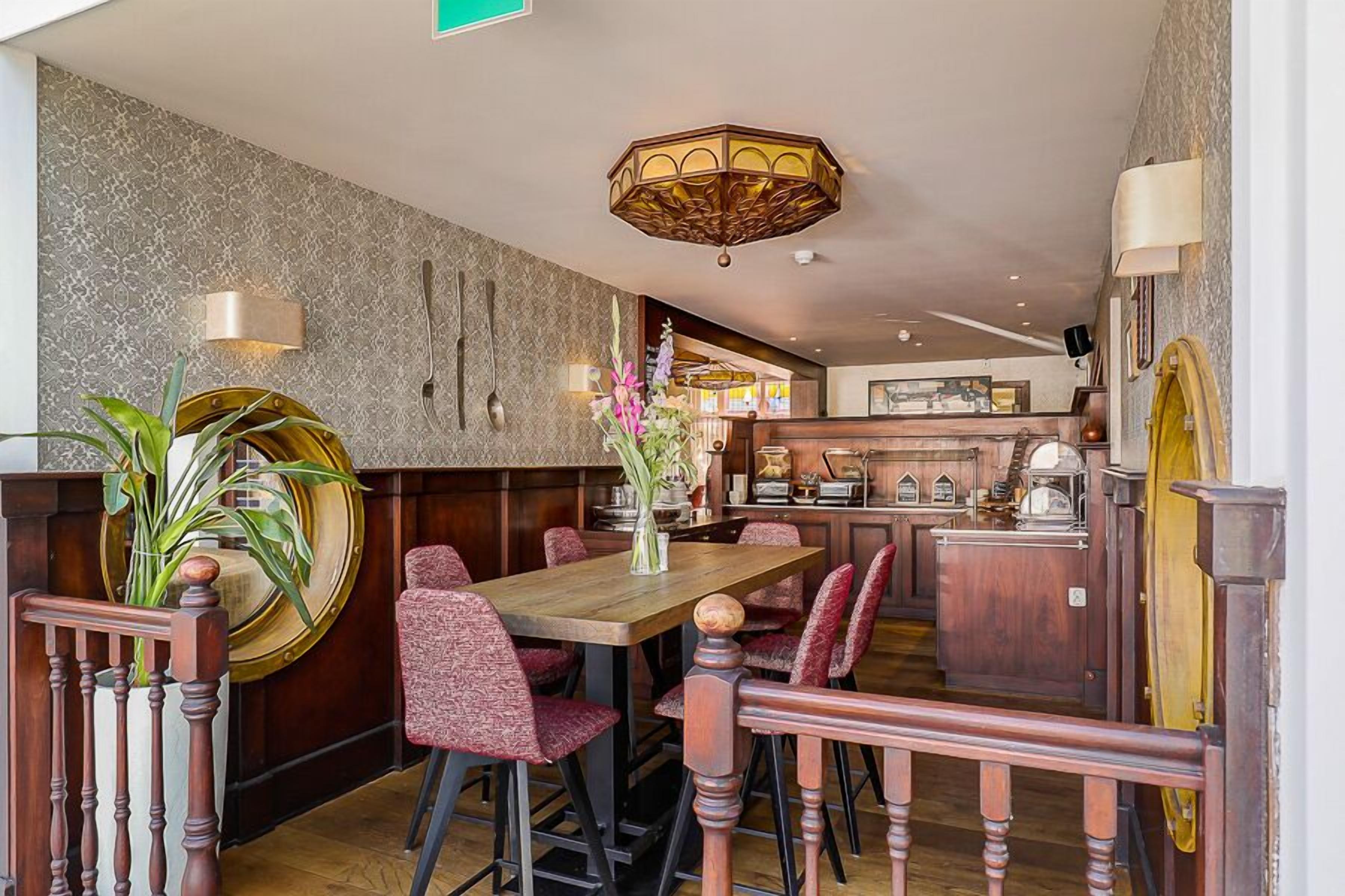 En restaurant med et trægulv, røde polstrede stole, et centralt bord med en vase med blomster, og et barområde i baggrunden.