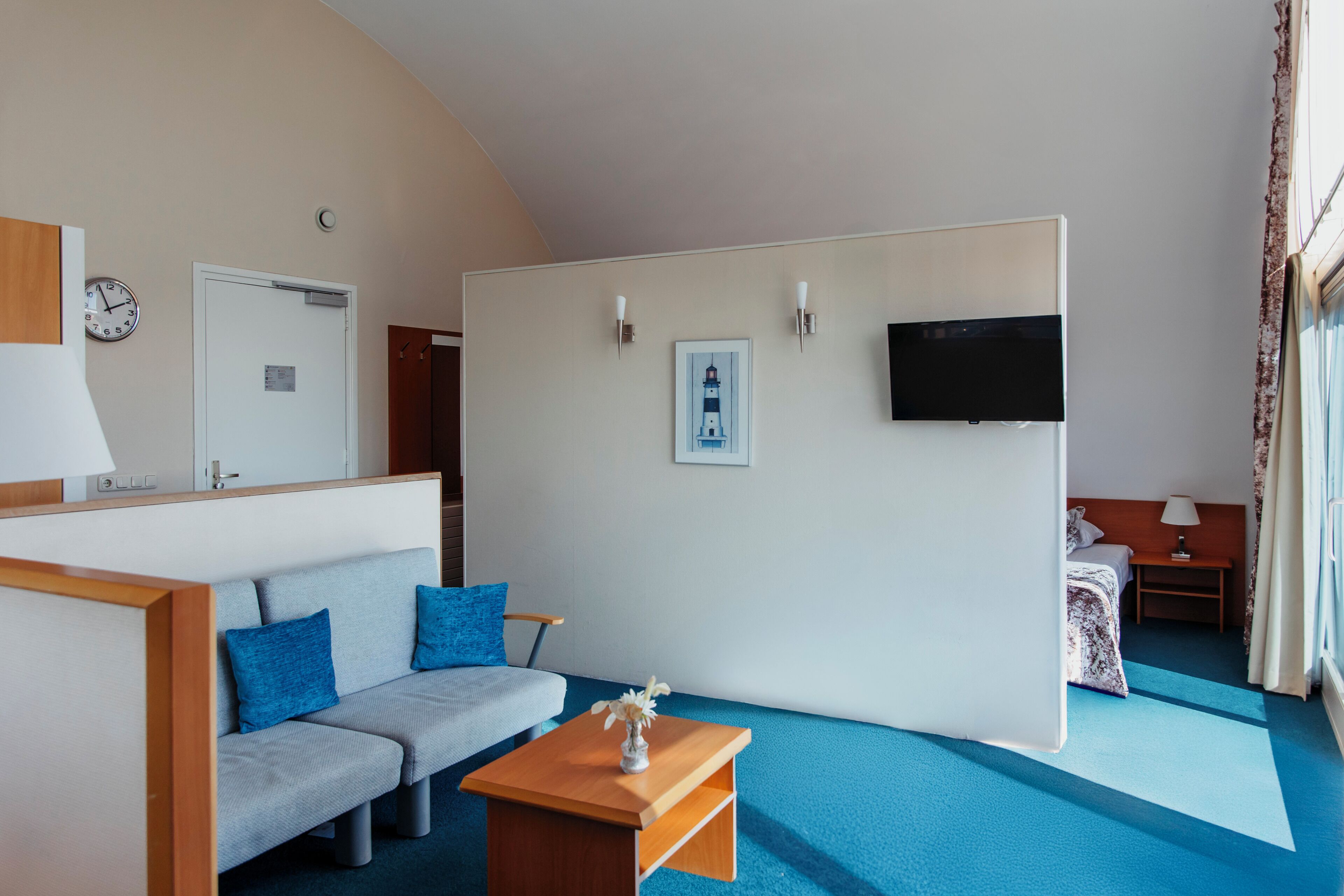 A hotel room with a sofa, a small table with a vase, a bed with a nightstand, a television mounted on the wall, and a door.
