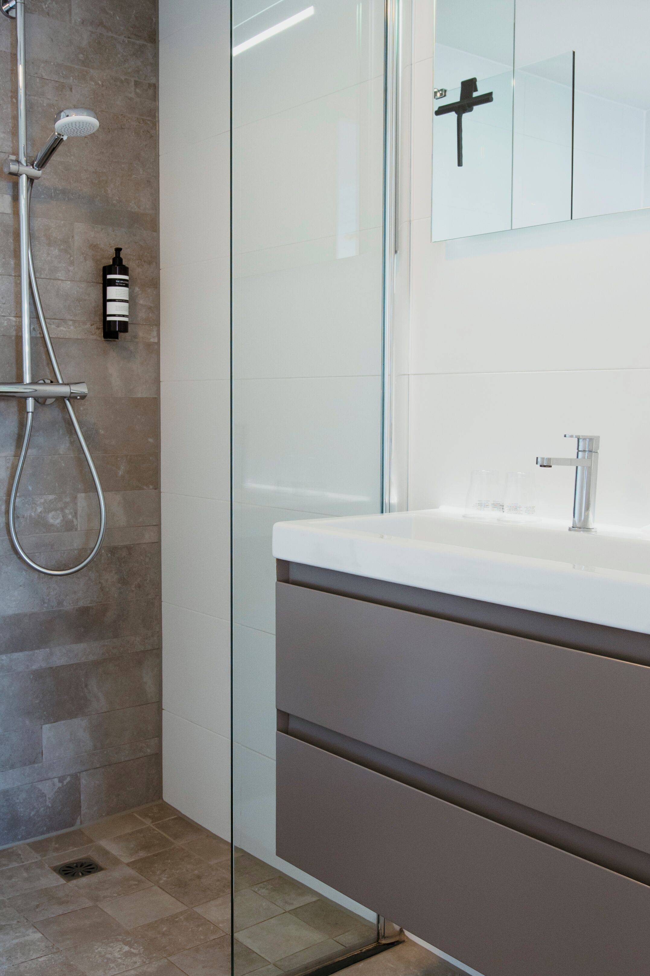 A modern bathroom with a glass shower enclosure, a white sink, and a mirror with a black cross.