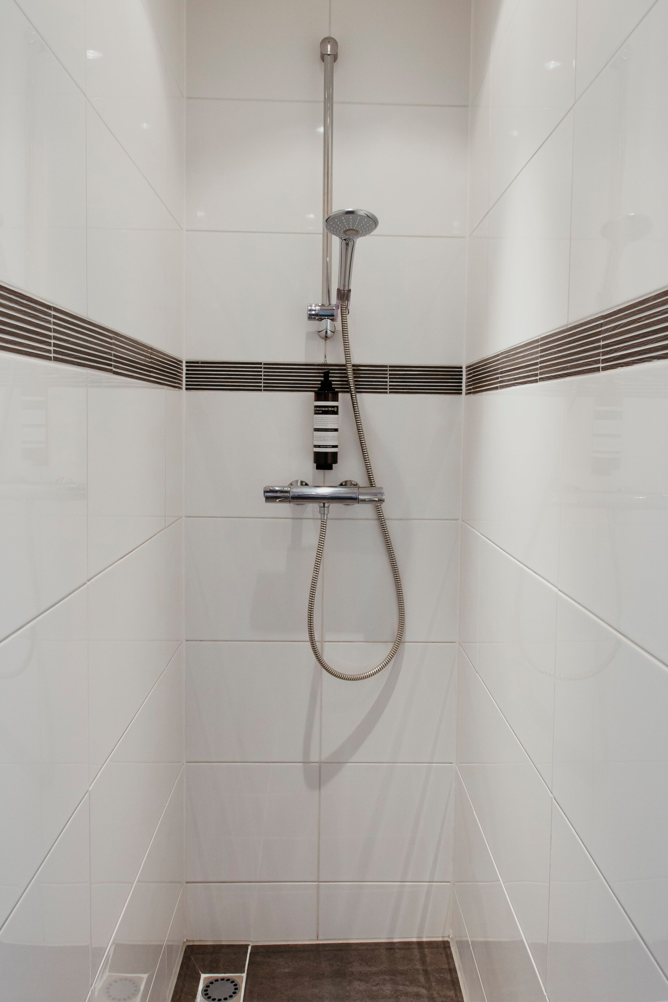 A modern shower with a handheld showerhead and a fixed wall-mounted showerhead, surrounded by white tiled walls with decorative black and brown tiles at the base.