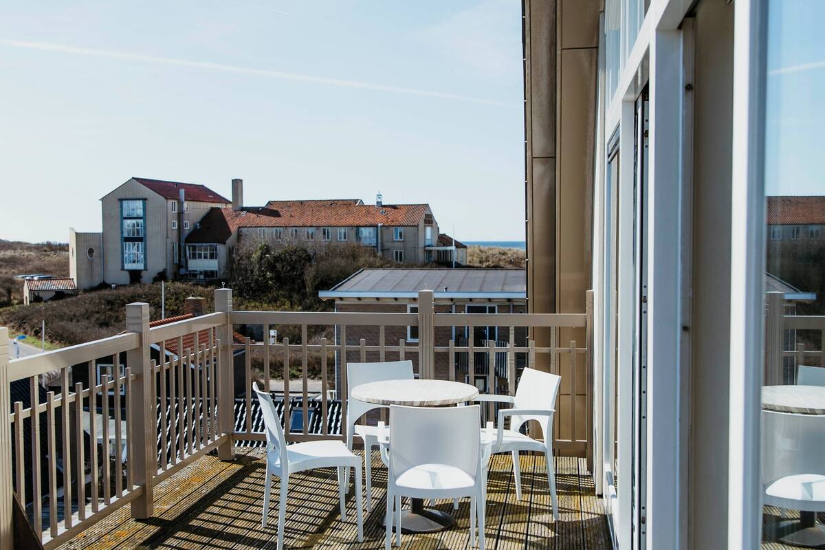 A wooden deck with white chairs and tables, overlooking a building with large windows and a view of the landscape.