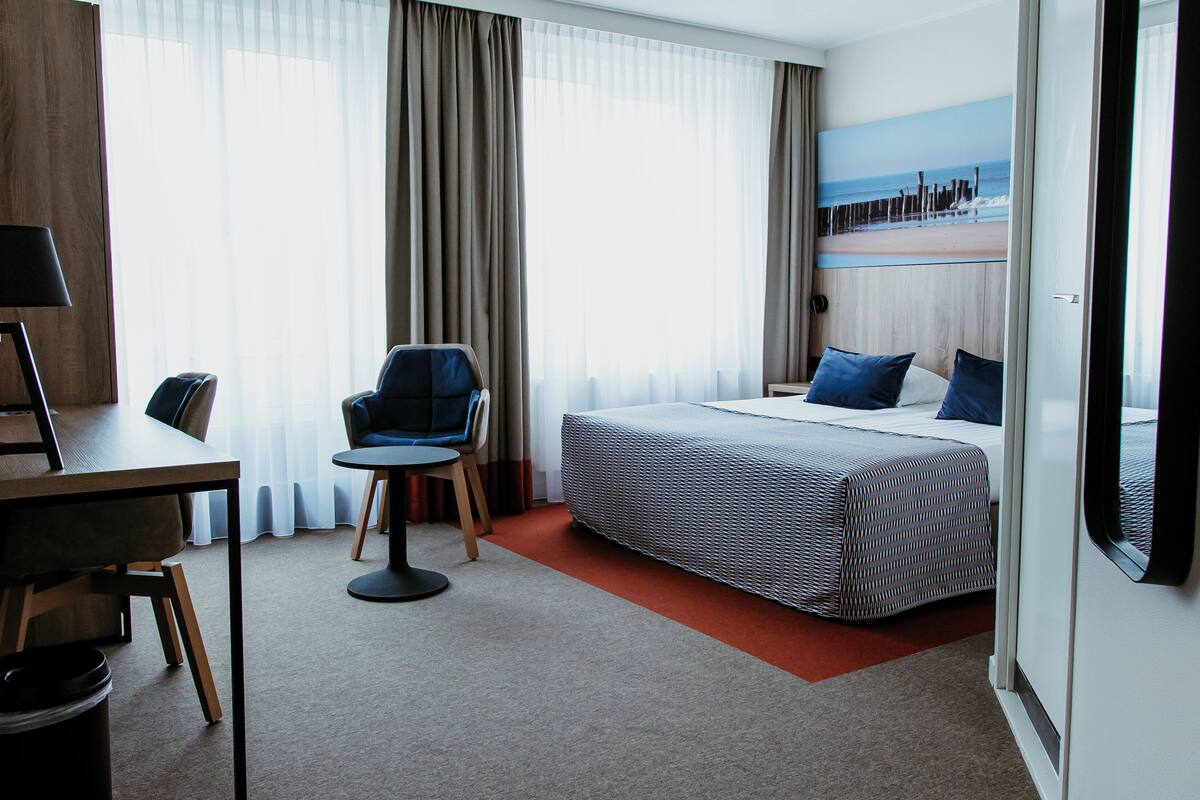 A hotel room with a bed, a desk, a chair, and a large window with curtains.