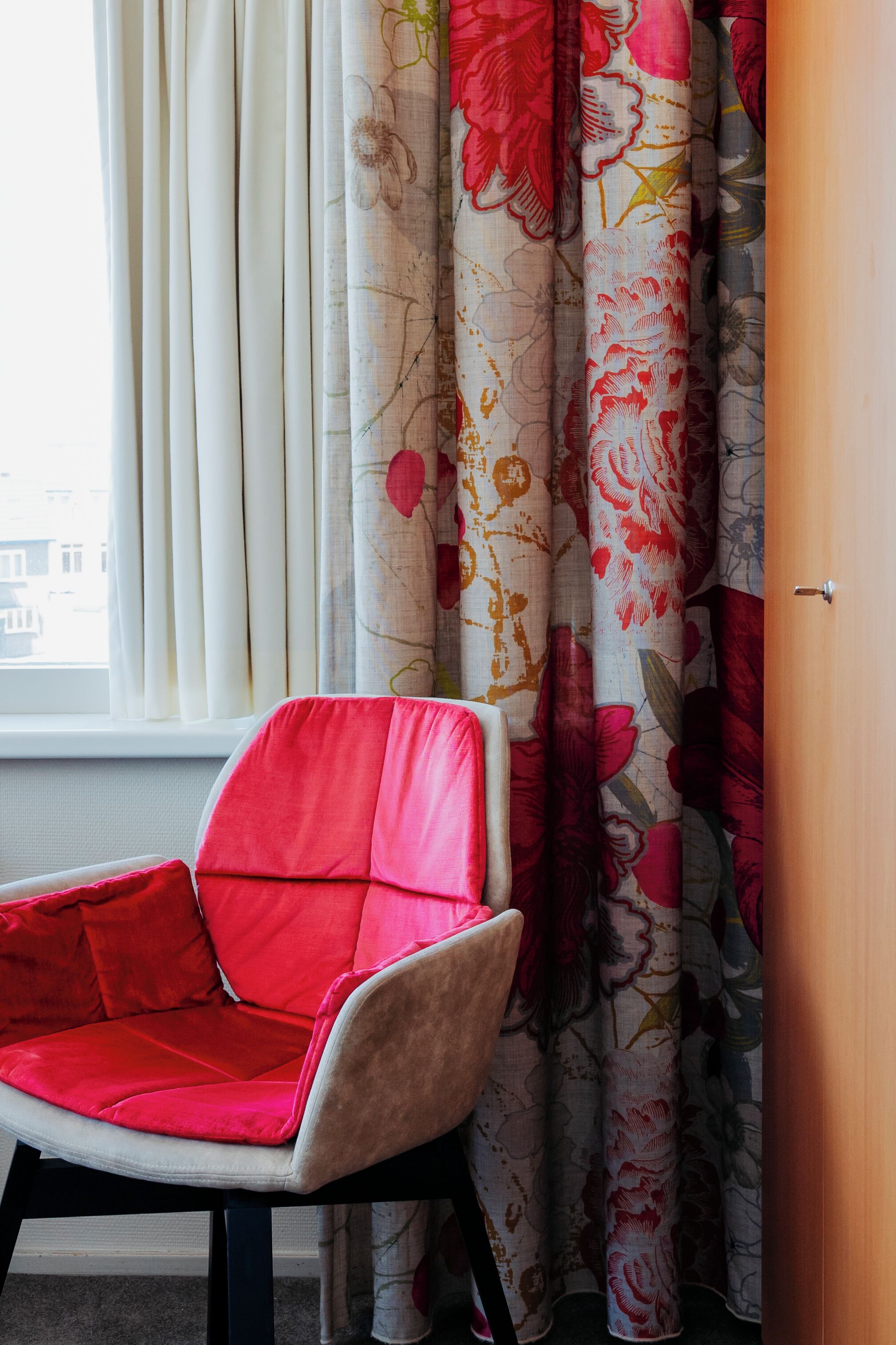 Una habitación con una silla roja, cortinas con motivos florales y una ventana que da a edificios.