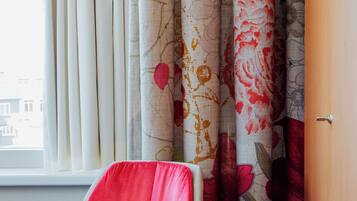 Una habitación con una silla roja, cortinas con motivos florales y una ventana que da a edificios.