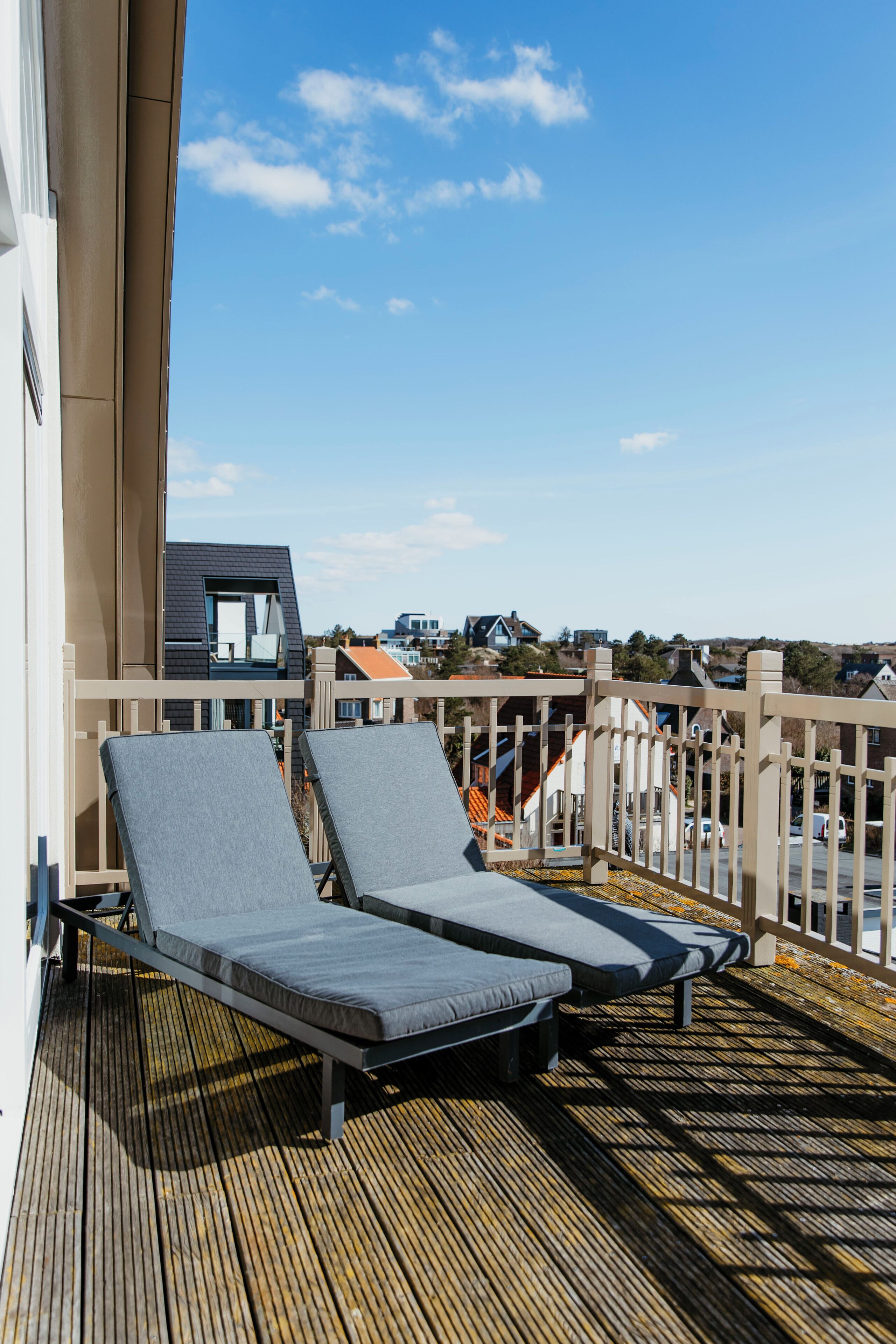 A balcony with two lounge chairs, a wooden deck, and a view of houses in the background.
