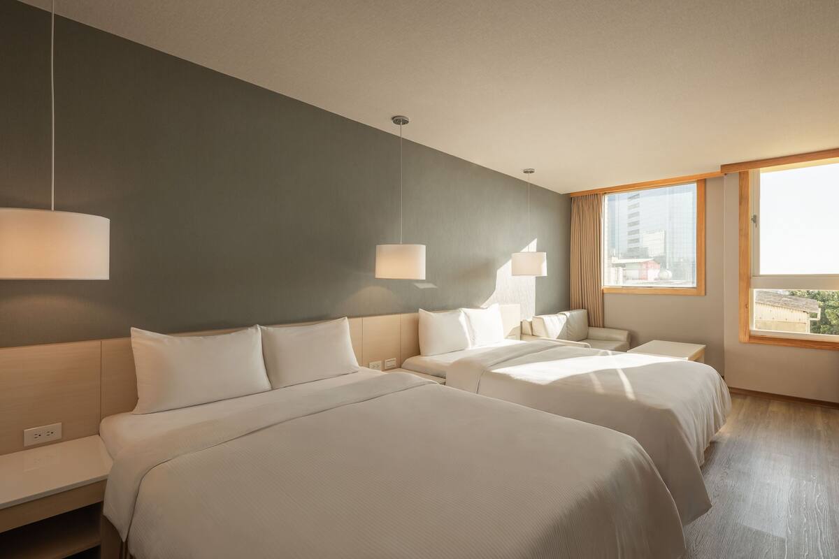 Habitación de hotel con dos camas, un ventanal grande y vistas a la ciudad.
