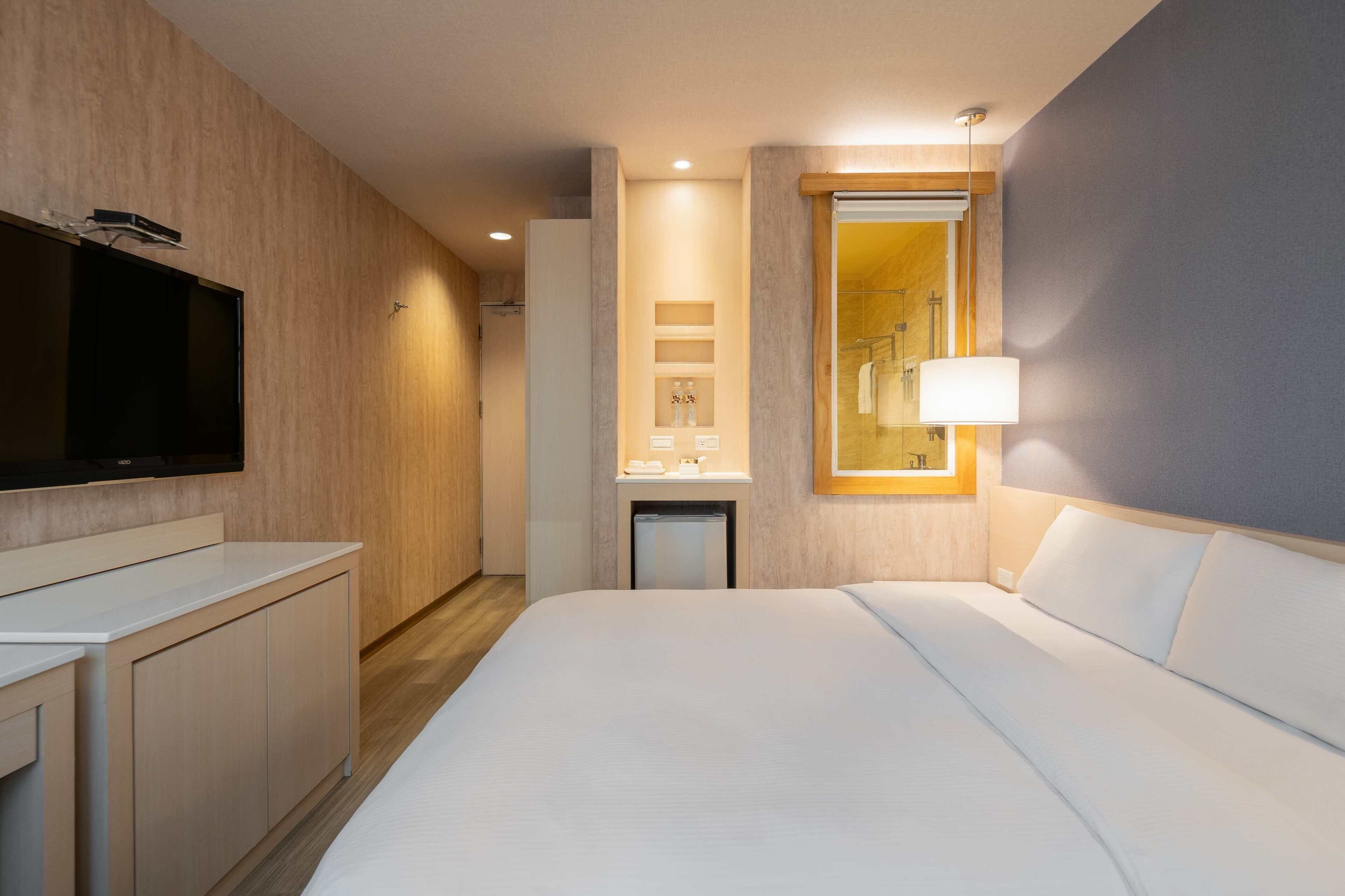 Habitación de hotel con cama, televisión montada en la pared, pequeño escritorio y espejo con lámpara.