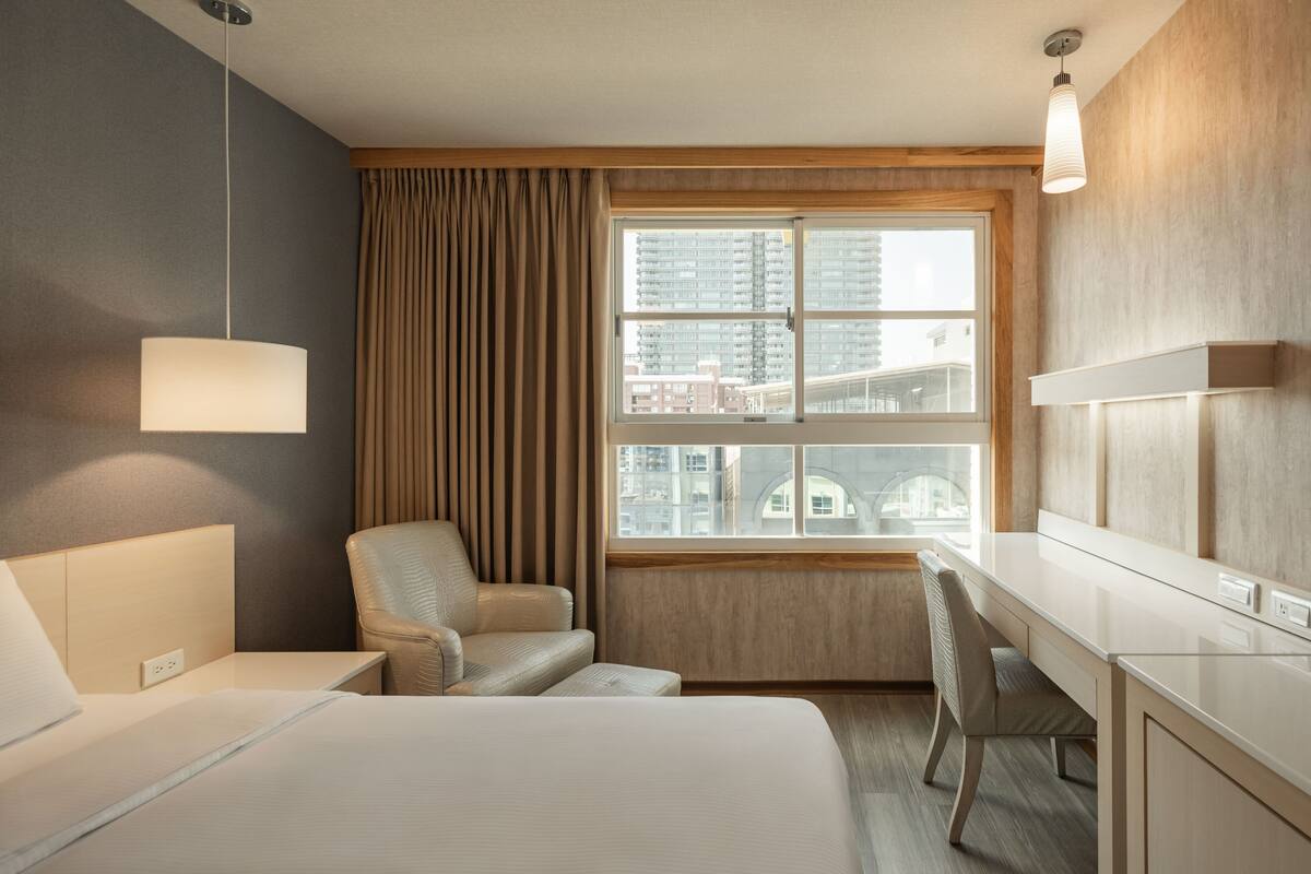 Habitación de hotel con cama, escritorio, silla, ventana con cortinas y lámpara colgante.