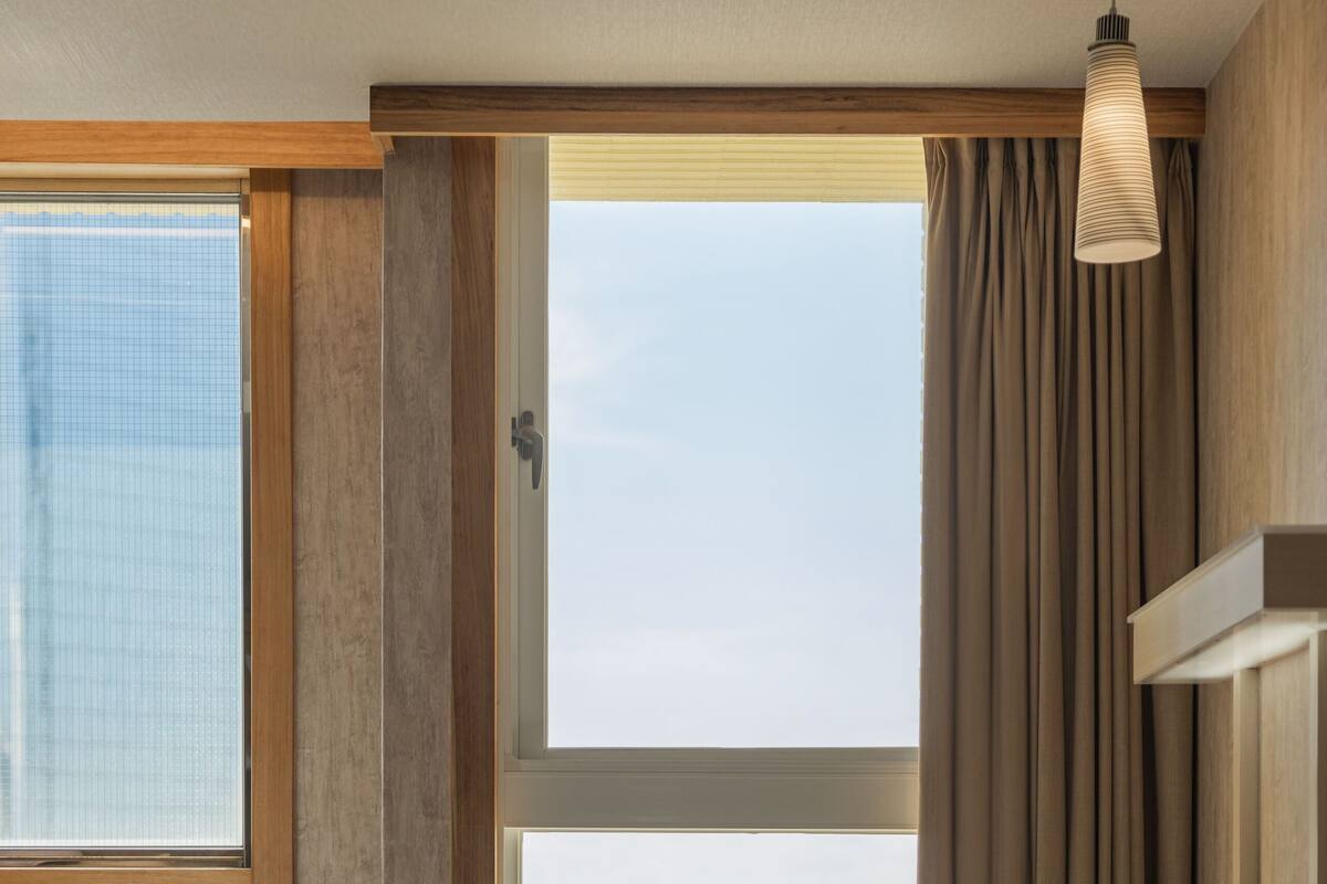 Habitación de hotel con cama, escritorio, silla y ventanas amplias que ofrecen vistas a la ciudad.