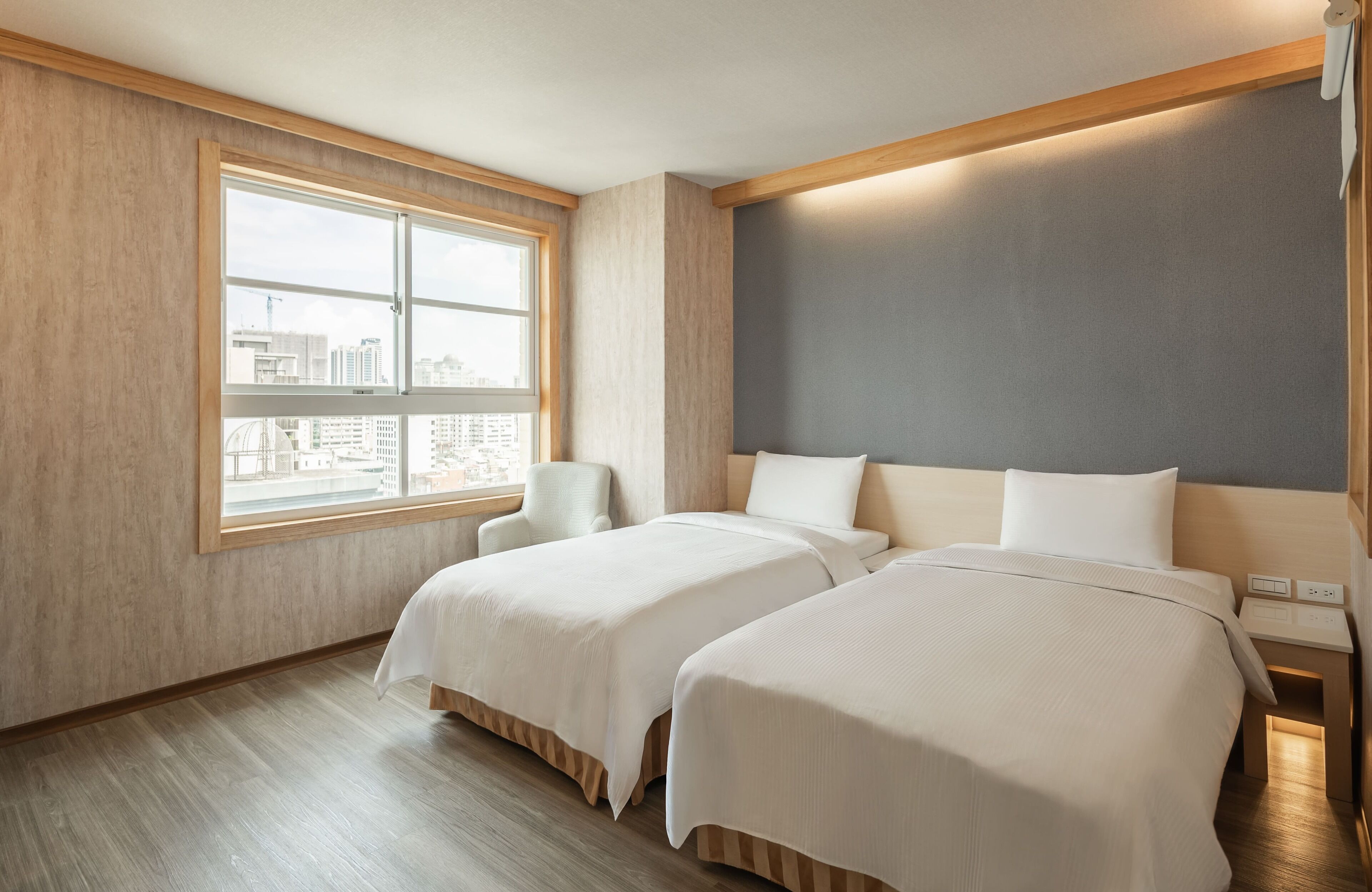 A hotel room with two beds, a large window with a city view, a chair, and a bedside table with a lamp.
