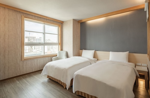 Habitación de hotel con dos camas, un ventanal grande con vistas a la ciudad, una silla y una mesita de noche con lámpara.