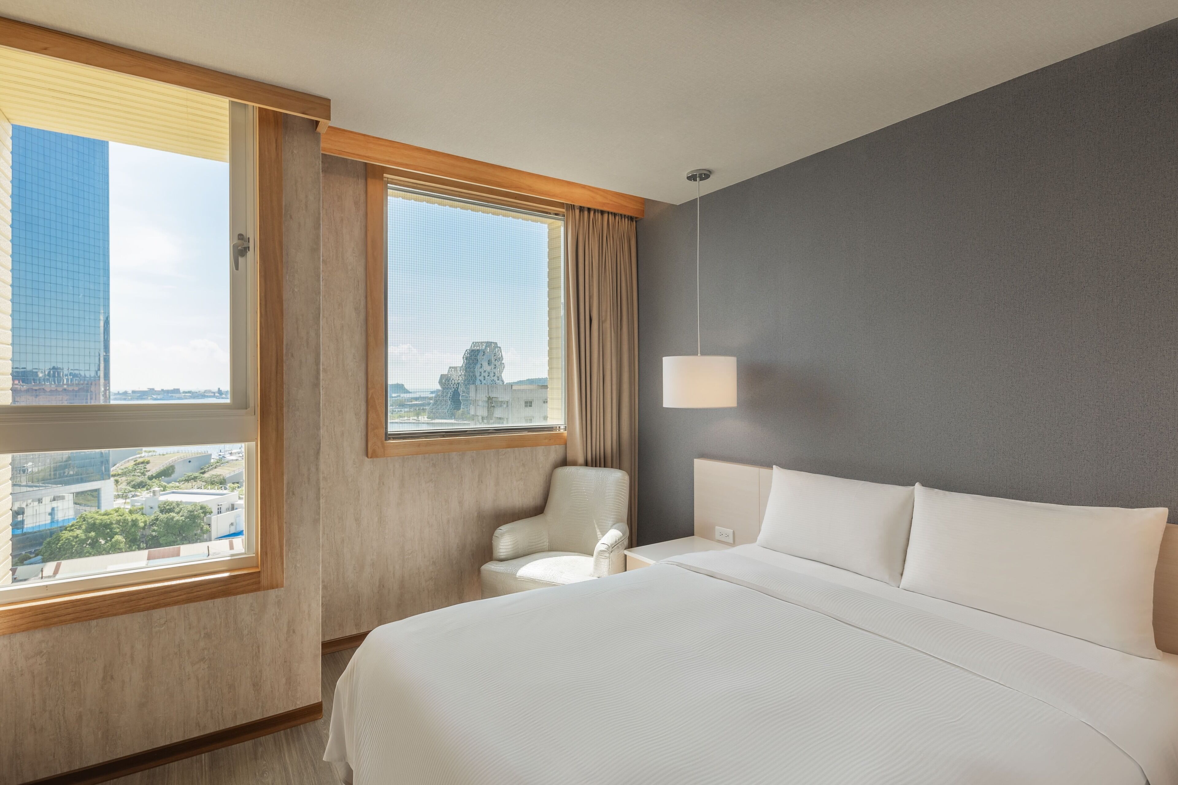 A hotel room with a large bed, a chair, a desk, and a window with a view of buildings.