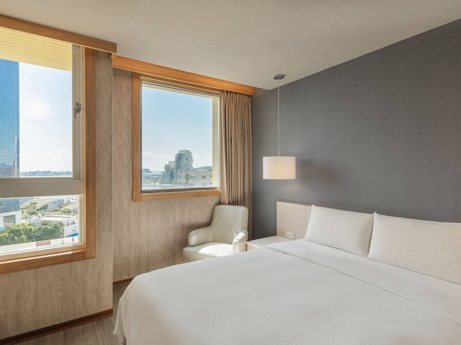 A hotel room with a large bed, a chair, a desk, and a window with a view of buildings.