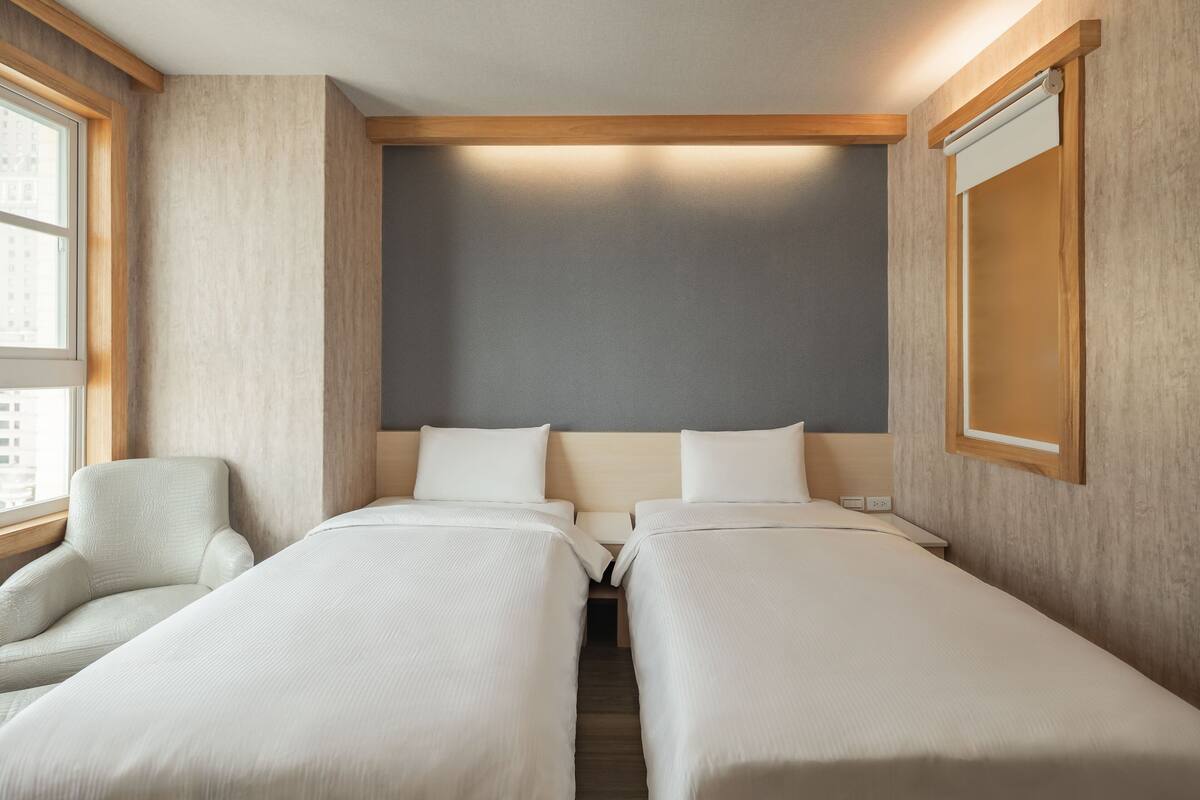 Habitación de hotel con dos camas individuales, una silla y un ventanal grande.