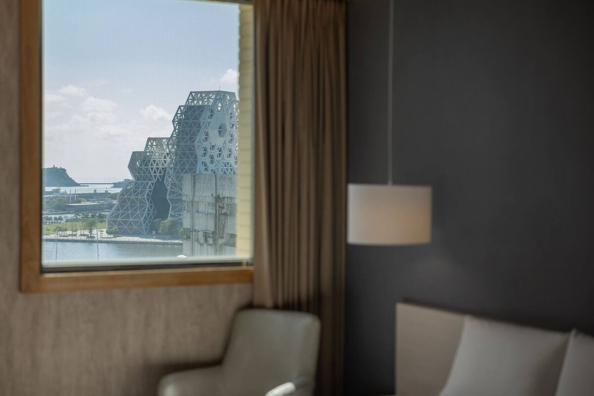 Habitación de hotel con un ventanal que ofrece vistas a un edificio moderno y una masa de agua.