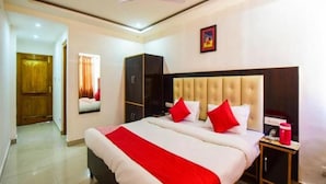 A hotel room with a large bed, a ceiling fan, a wardrobe, and a small table with a red object on it. - Anand Palace Hotel (Udaipur)