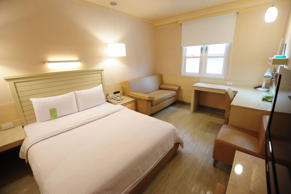 Ein Hotelzimmer mit Bett, Nachttisch, Lampe, Schreibtisch, Stuhl und einem Fenster.