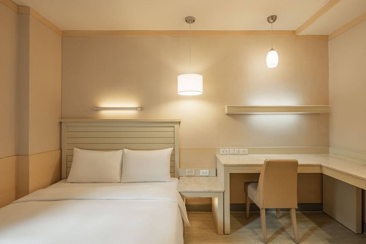 Ein Hotelzimmer mit einem Bett, einem Schreibtisch und einem Sessel. Zwei Wandleuchten sind angebracht.