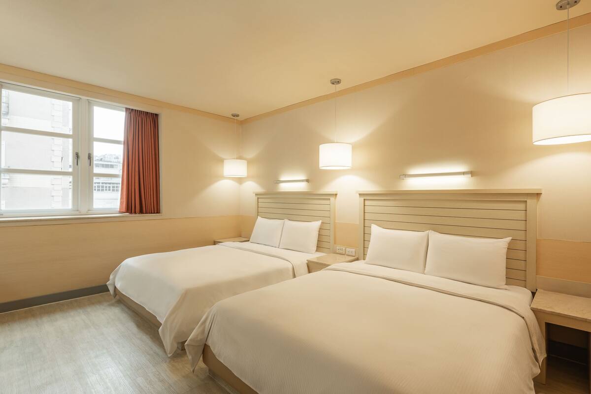 Ein Hotelzimmer mit zwei Betten, einem fensterbelaubten Fenster und Wandleuchten.