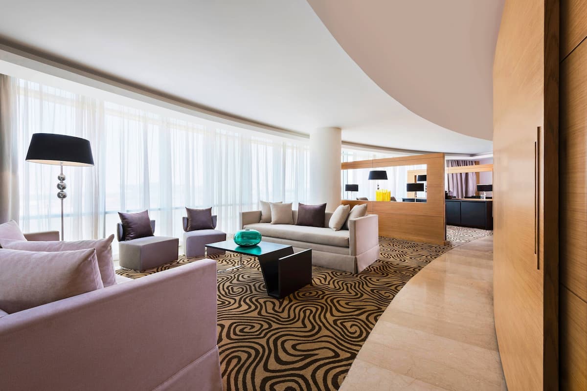 Un hall d’hôtel moderne avec un mur en bois aux lignes courbes, un espace salon avec des canapés et des fauteuils, une moquette à motifs et une grande fenêtre ornée de rideaux légers.